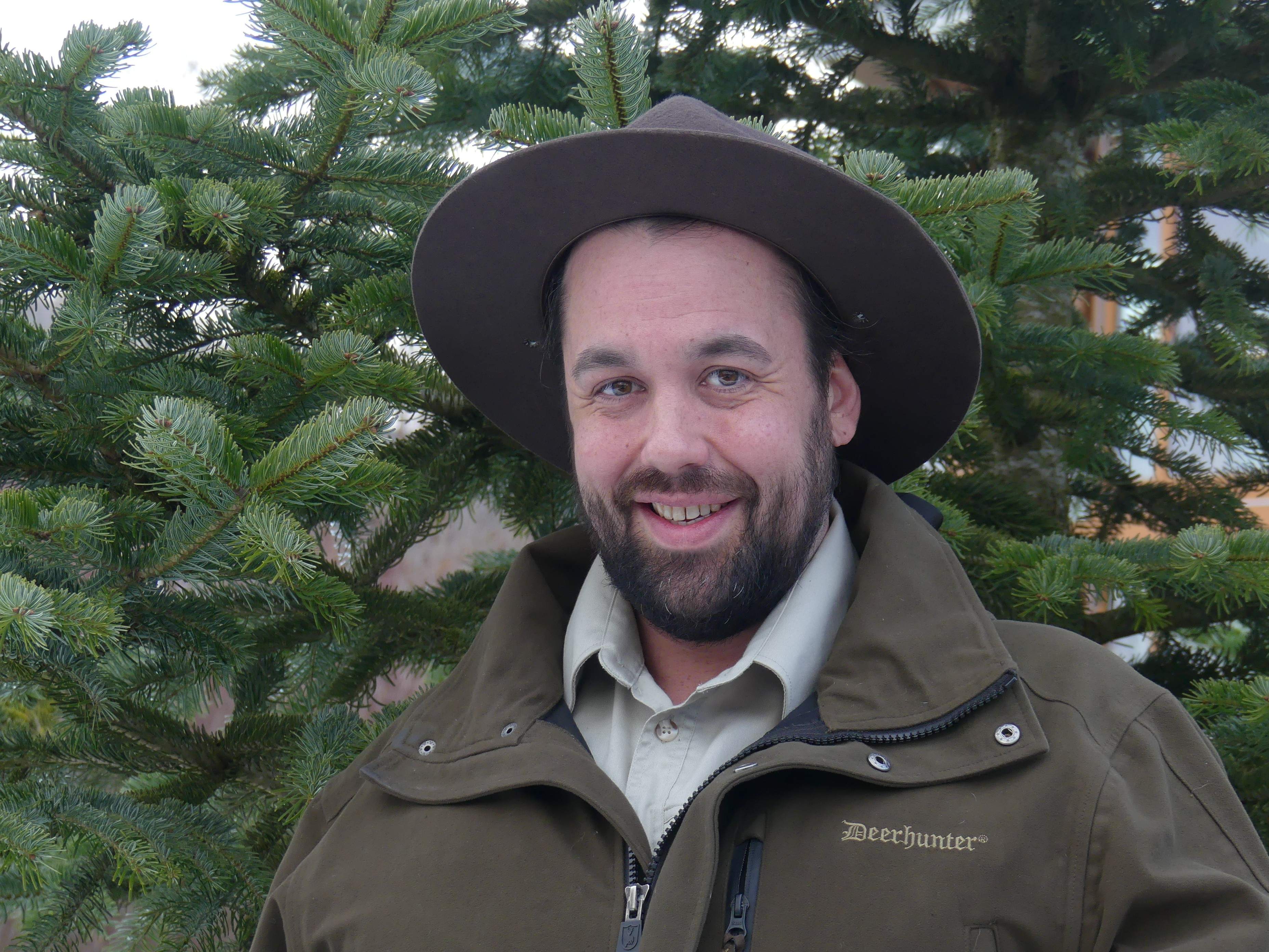 Der Ranger steht vor einem Nadelbaum und lächelt in die Kamera. Er hat einen Rangerhut auf.