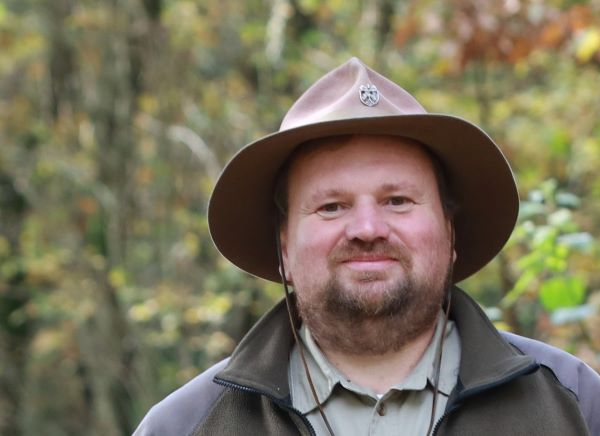 Der Ranger steht vor belaubten Bäumen und lächelt in die Kamera. Er hat einen Rangerhut auf.