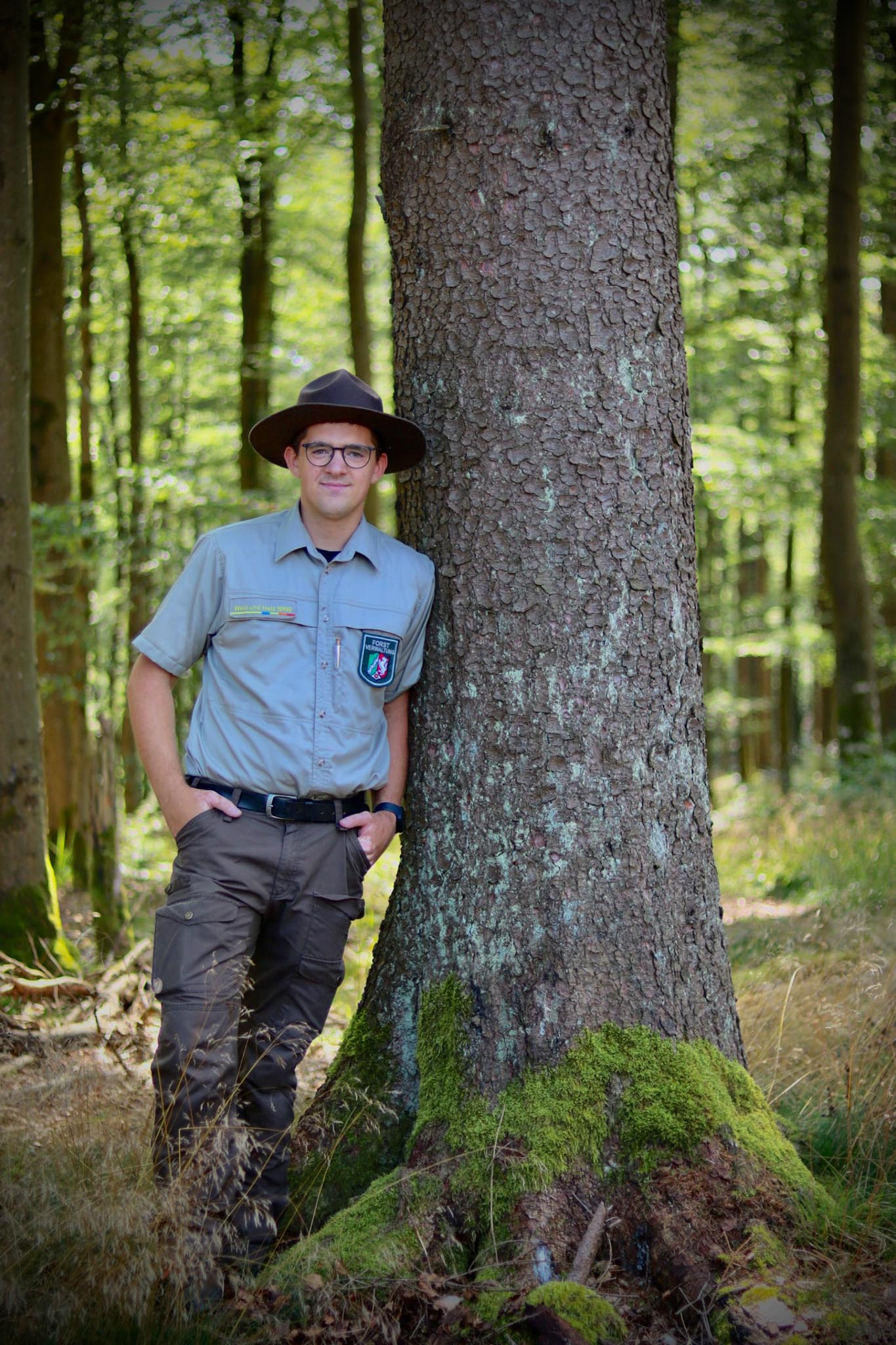 Ein Ranger steht links neben einem dicken Baum im Wald. Er lehnt mit seiner Schulter an den Stamm. Er hat einen Rangerhut auf und lächelt in die Kamera.
