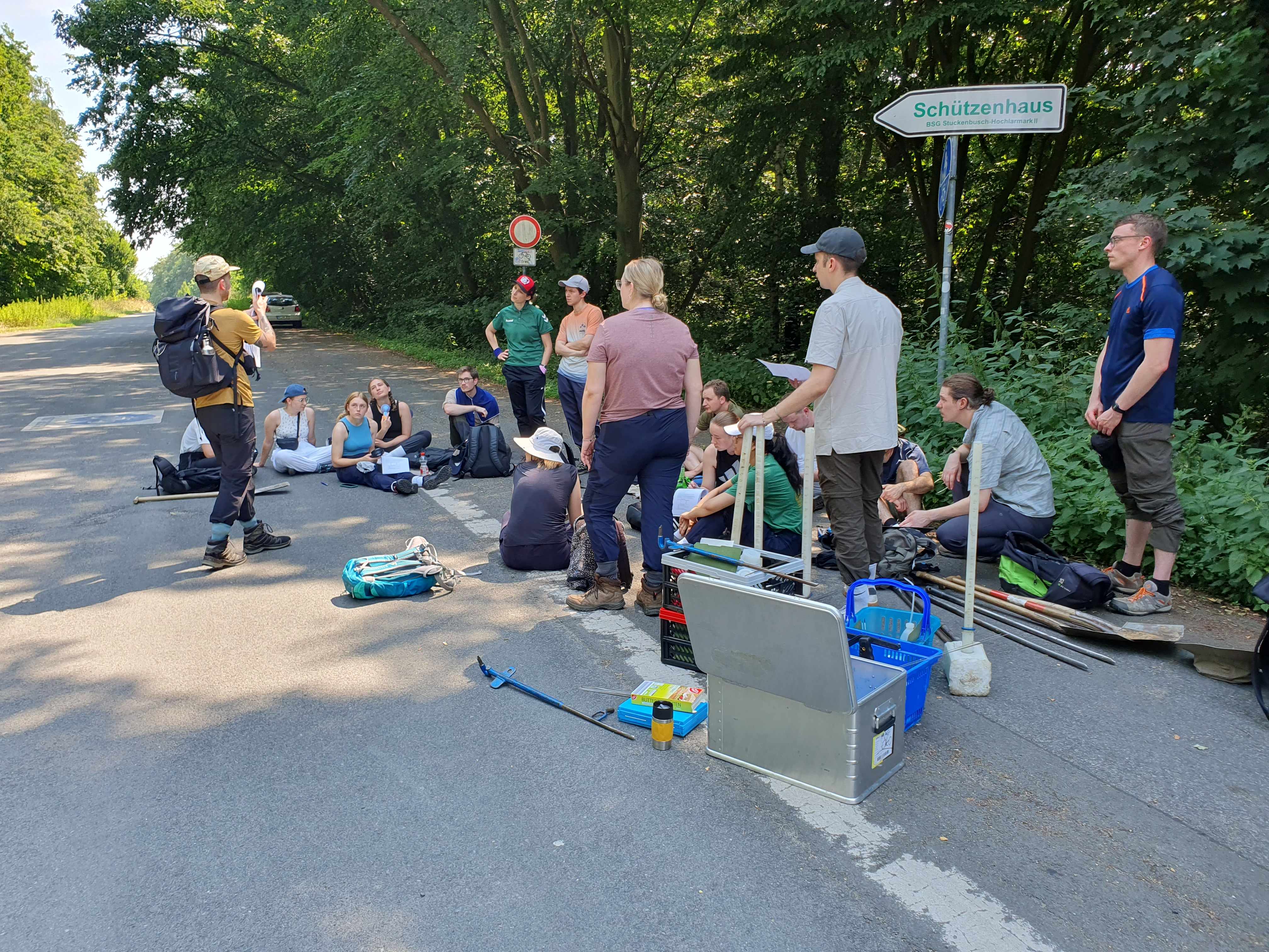 Die Studenten stehen und sitzen auf einer Straße am Waldrand. Um sie herum stehen Boxen und Werkzeuge 
